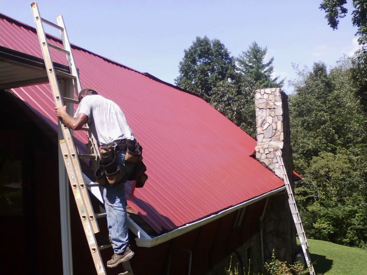 Roofer on a ladder beside a red metal roof and stone chimney — Soffit & Fascia Repair by Roofing Brothers in Russellton
